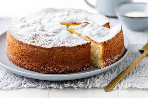 Una soffice torta allo yogurt, cosparsa di zucchero a velo, attende di essere gustata. Una fetta è già stata tagliata, rivelando il suo interno invitante.
