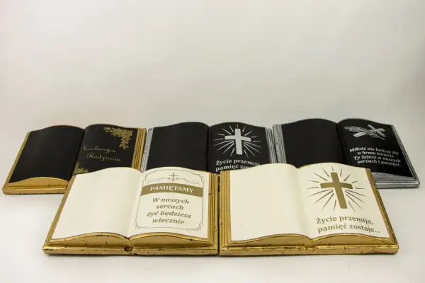 Otwarta książka na cmentarz, złota, z napisem "Pamiętamy" i krzyżem. Obok inne nagrobki w kształcie ksiąg.