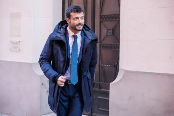 Un homme élégant porte un type de manteau homme bleu marine, un costume et une cravate, marchant dans la rue.