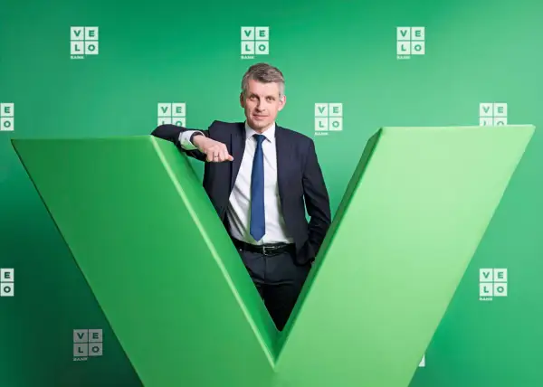 Mężczyzna w garniturze opiera się o zieloną literę "V" z logo Velo Bank.