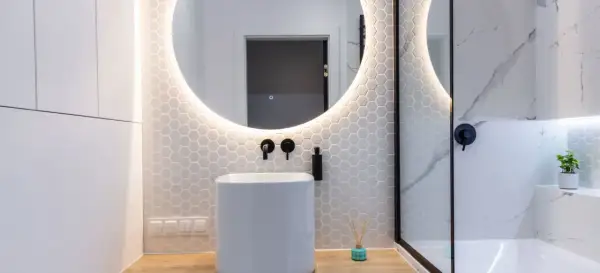 Moderno baño con lavabo blanco, espejo redondo iluminado y azulejos hexagonales. Ideal para tu presupuesto baño.