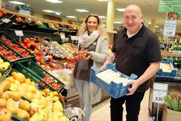 Mann und Frau im Markt Refrath mit frischem Obst und Gemüse. Er hält eine Kiste Spargel, sie Erdbeeren.