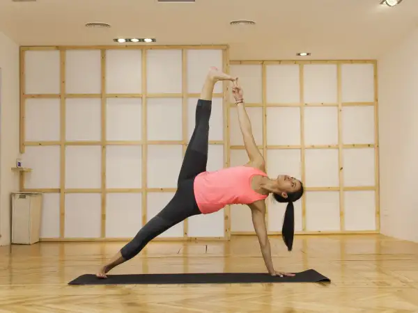 Mujer en postura de plancha lateral yoga, con pierna elevada y mano extendida hacia el techo.