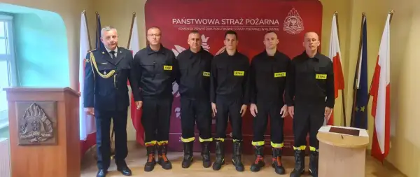 Sześciu strażaków w mundurach, w tym dowódca, tworzy rota strażacka. Stoją na tle sztandarów i logo Państwowej Straży Pożarnej.