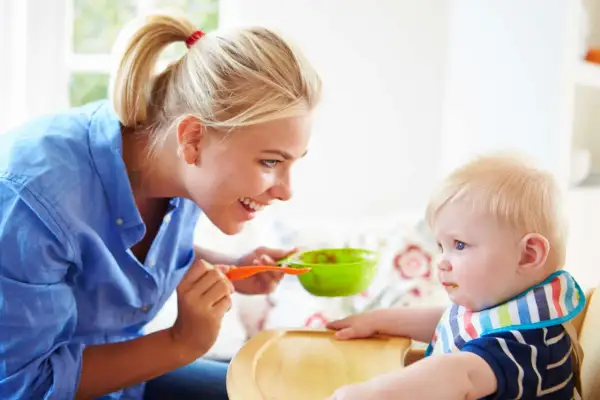 Mama karmi dziecko łyżeczką z zielonej miseczki. Dziecko ma śliniaczek, a mama uśmiecha się do niego. To posiłek bogaty w błonnik dla dziecka.