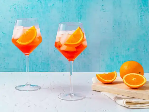 Två glas med Aperol Spritz, is och apelsinskivor, med en hel apelsin och skivor på en skärbräda i bakgrunden.