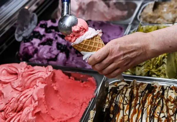 Eine Hand füllt eine Waffel mit rosa Eis. Daneben stehen Behälter mit verschiedenen Eissorten, darunter Blaubeer- und Pistazieneis. Das beste Eis der Welt!