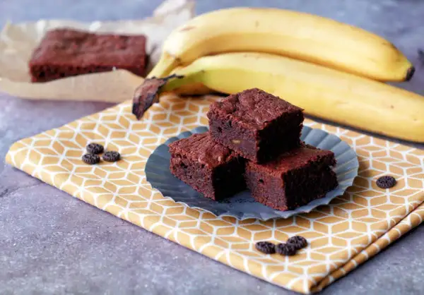 Saftige Bananen-Brownies auf einem gelben Tuch, daneben reife Bananen. Ein Genuss!