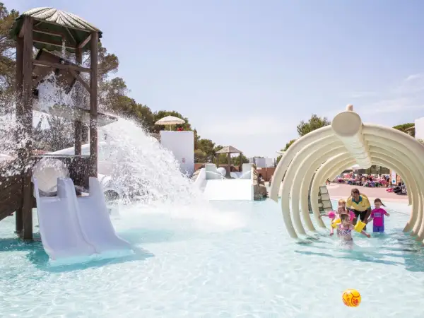 Kinder planschen im Wasserpark, während ein Wasserfall vom Rutschenturm herabstürzt. Ein Mann mit gelbem Shirt beaufsichtigt die Kleinen unter einem knochenartigen Tunnel in den Calimera Fido Gardens.