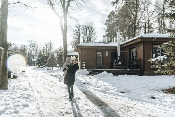Kobieta z dzieckiem na rękach idzie zimową drogą do drewnianego domku.