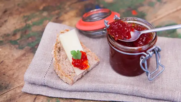 Un cucchiaino raccoglie la lucida marmellata di peperoni da un barattolo. Sul pane, formaggio e un tocco di questa delizia.