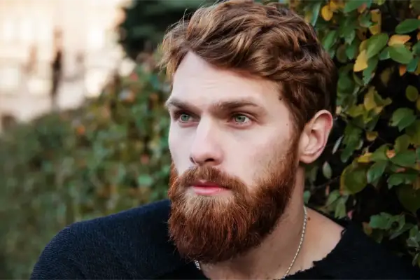 Un homme au visage ovale, avec une barbe rousse et des yeux bleus perçants, regarde au loin, entouré de verdure.