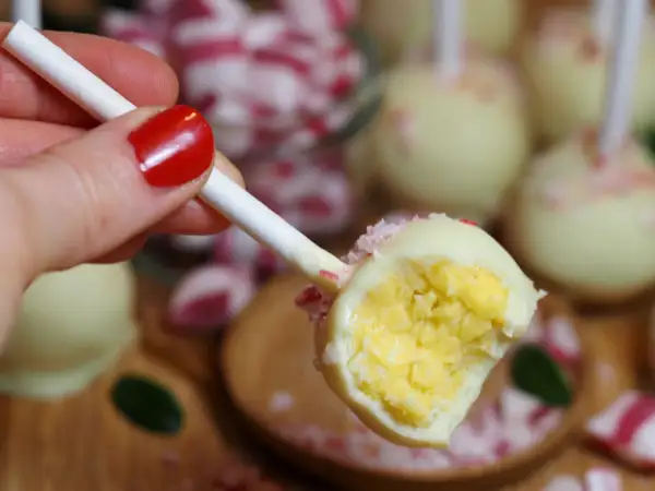 En hand med rött nagellack håller en cake pop med vit choklad och krossade polkagrisar. Perfekt för ett cake pops recept!