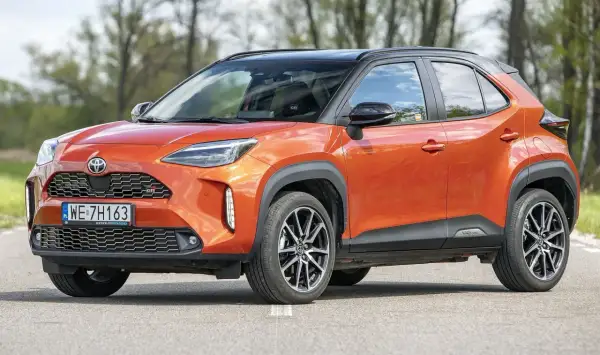Pomarańczowy Toyota Yaris Cross 2024 z czarnym dachem, zaparkowany na drodze, z drzewami w tle.