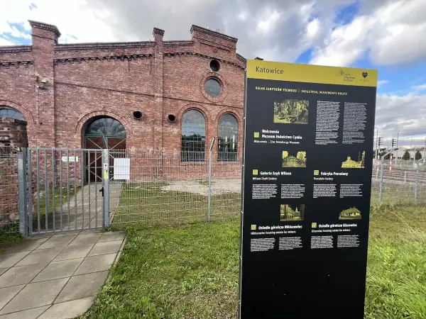 Tablica informacyjna Szlaku Zabytków Techniki w Katowicach, obok ceglanej budowli dawnej huty.