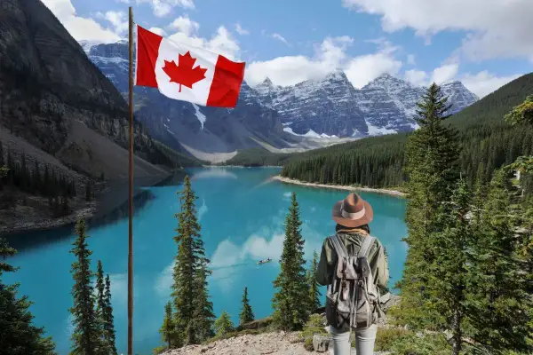 Persona con mochila admira el lago turquesa y las montañas nevadas de Canadá. La bandera canadiense ondea, invitando a una aventura de work and holiday Canada.