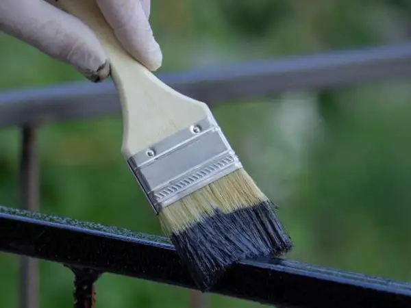 Mano con guanto bianco che usa un pennello per applicare vernice scura su una ringhiera, un passo per come sverniciare una ringhiera.