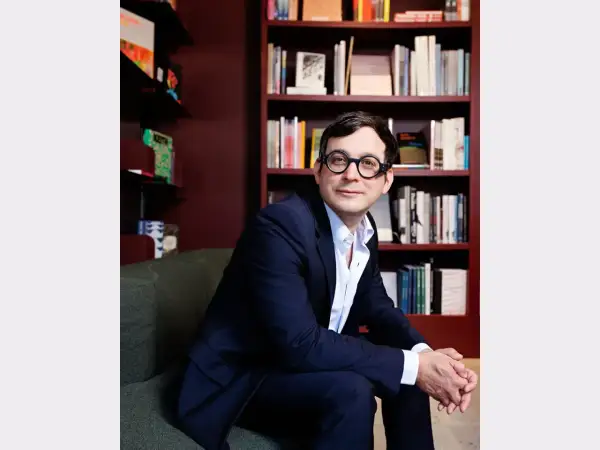 A man in a suit and glasses sits in front of bookshelves, perhaps at the David Lewis Gallery, looking thoughtfully to the side.
