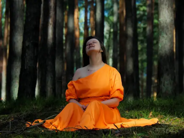 Kobieta w pomarańczowej sukni siedzi na trawie w lesie, ciesząc się chwilą kontaktu z naturą.