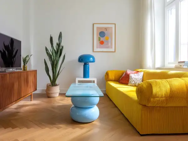 Jasny salon retro z żółtą kanapą, niebieską lampą i stolikiem kawowym. Drewniana komoda i duża roślina dopełniają przytulną atmosferę.