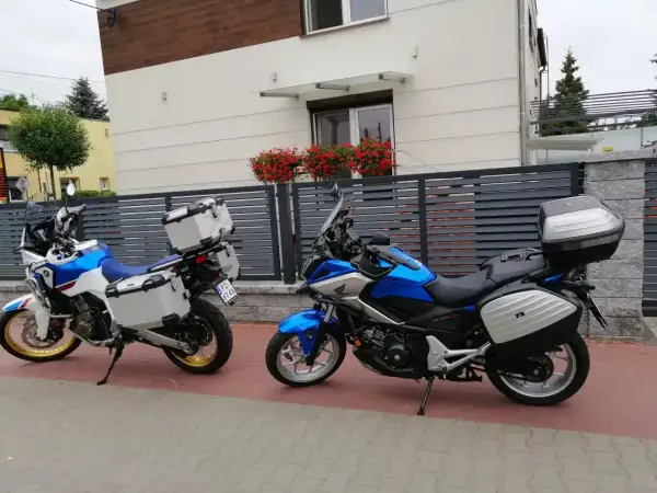 Dwa motocykle turystyczne z kuframi stoją przed nowoczesnym domem z kwiatami na balkonie.