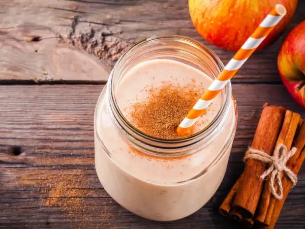 Pyszny drink szarlotka w słoiku, posypany cynamonem, obok jabłka i laski cynamonu na drewnianym tle.