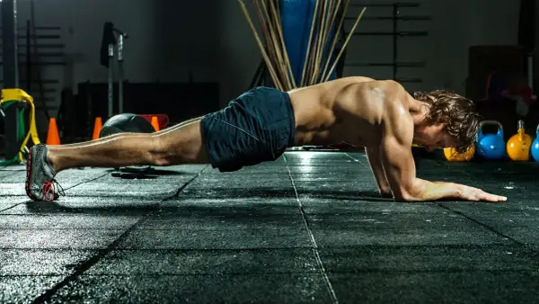 Mann in Unterarmstützposition, um zu zeigen, wie oft bauchmuskeln trainieren. Im Hintergrund sind Kettlebells und Trainingsgeräte.