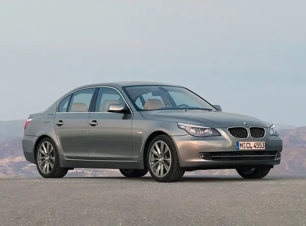 Szary sedan BMW 5 Series (E60) z jasnym wnętrzem stoi na pustej drodze, z górami w tle.