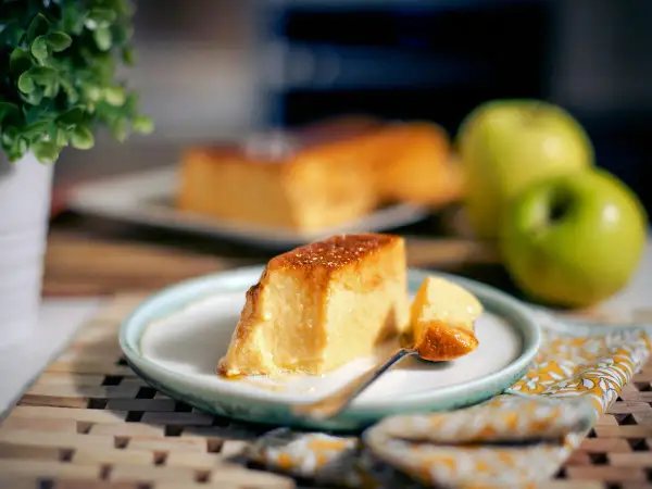 Un delicioso flan de manzana, con una porción servida en plato, junto a manzanas verdes y una planta.
