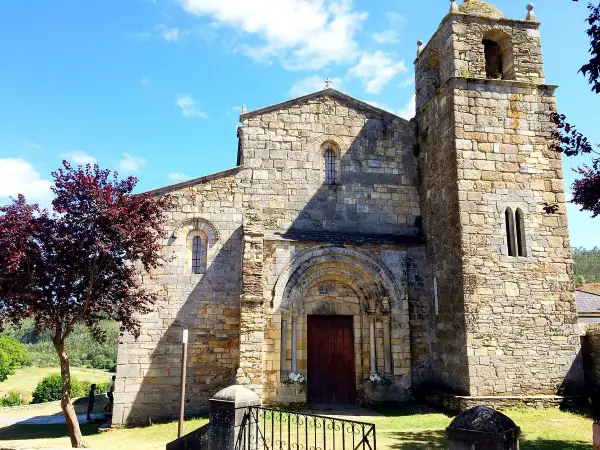 La catedral de Foz, con su torre románica y fachada de piedra, se alza bajo un cielo azul con nubes. Un árbol de hojas rojizas añade color.