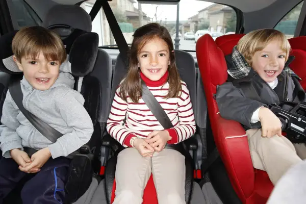Tres niños sonríen en sus asientos de coche. El niño de la izquierda, con sudadera gris, parece preguntarse cuándo puede ir un niño delante.