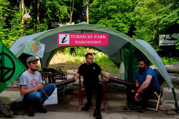 Trzech mężczyzn siedzi pod namiotem z napisem "Turnicki Park Narodowy". Obok zielona flaga z symbolem.