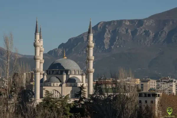 Imponujący meczet z minaretami, górujący nad budynkami stolicy Albanii, na tle skalistych gór.