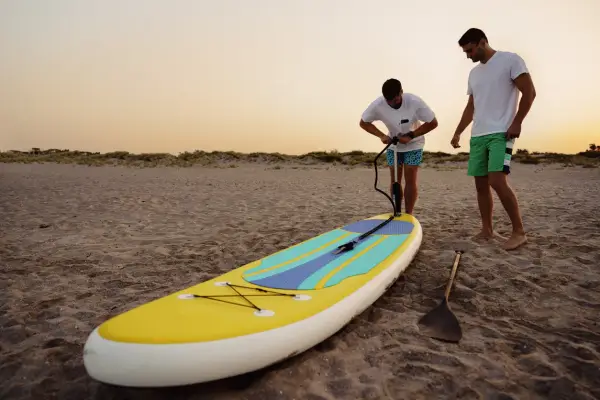 Zwei Männer bereiten ein SUP-Board am Strand vor. Einer pumpt es auf, fragt sich: "Sup, wieviel Bar?
