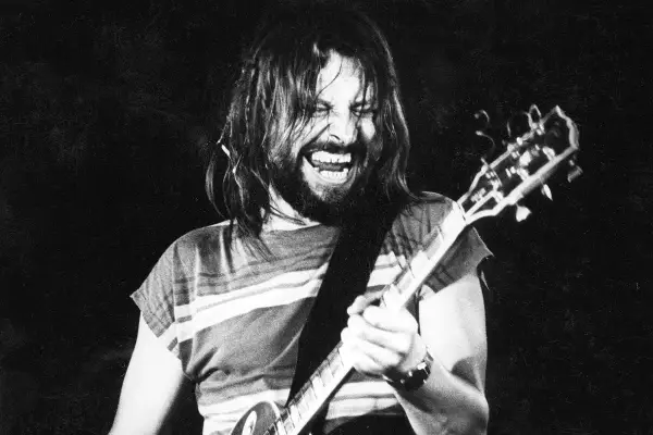 Rockman z długimi włosami i brodą, grający na gitarze z pasją. Jego twarz wyraża emocje, jakby to była jego osobista dżem modlitwa.