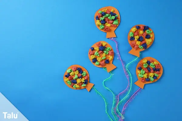 Bunte Ballons aus Papier, ideal zum Fasching Deko basteln. Kleine, zerknüllte Papierstücke in Rot, Blau, Grün und Gelb zieren die orangefarbenen Ballons.