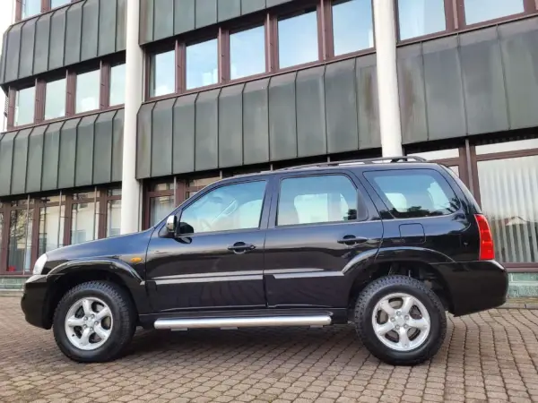 Czarny Mazda Tribute stoi na brukowanym podjeździe przed nowoczesnym budynkiem.