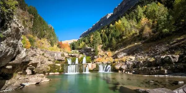 Cascada en un valle montañoso, ideal para rutas senderismo España. El agua verde turquesa fluye entre rocas y árboles otoñales.