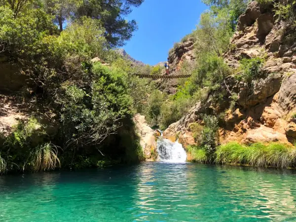 Un puente colgante cruza un **río verde** en Granada, con una cascada y vegetación exuberante.