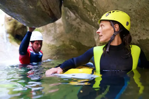 Kobieta w żółtym kasku i piankowym stroju zanurzona w wodzie, obok mężczyzna w białym kasku. To właśnie canyoning – przygoda w kanionie.