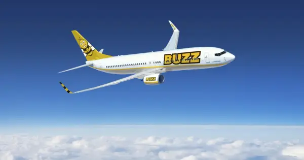 Samolot Buzz, należący do Ryanair, leci nad chmurami. Słońce odbija się od białego kadłuba, a na ogonie widnieje uśmiechnięta pszczoła. Idealny na wakacje z bagażem!