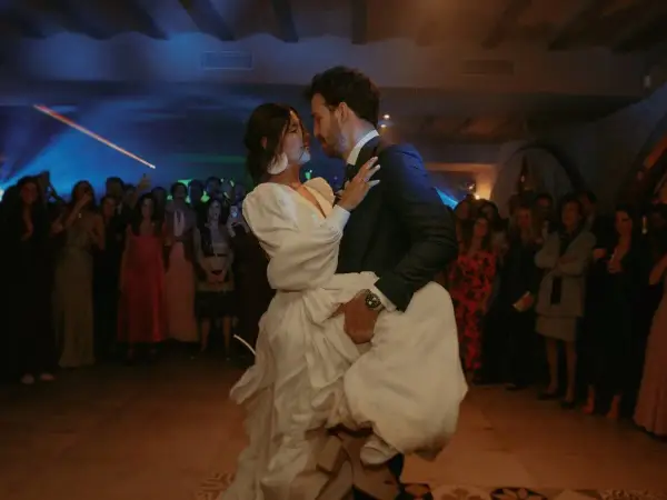 Novios bailando su vals boda, rodeados de invitados expectantes. La novia, con un vestido blanco voluminoso, es alzada por el novio.