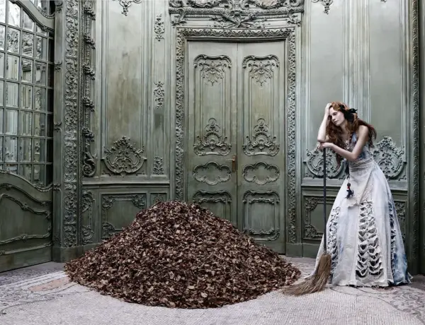 A woman in a tattered gown leans on a broom beside a mountain of leaves in a grand, ornate room, a scene captured by Eugenio Recuenco.