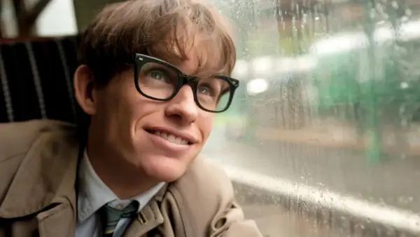 Junger Stephen Hawking mit Brille blickt lächelnd aus einem Fenster, das von Regentropfen bedeckt ist.