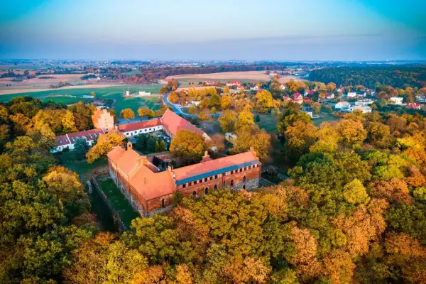 Zamek Bierzgłowski otoczony jesiennym lasem, z czerwonymi dachami i ceglanymi murami, w blasku zachodzącego słońca.