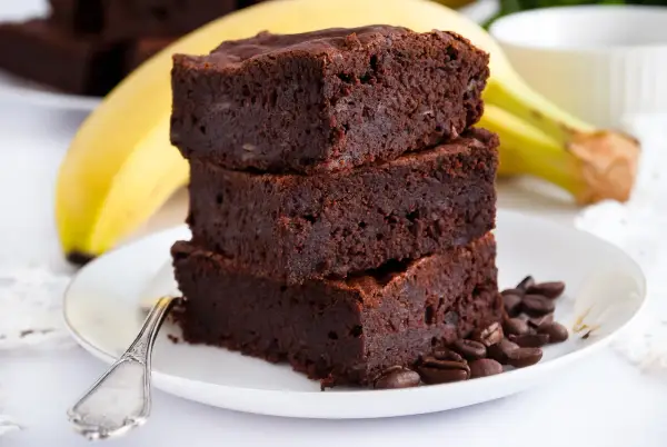 Stos puszystych brownie z bananami, ozdobiony ziarnami kawy, na talerzu z bananem w tle.