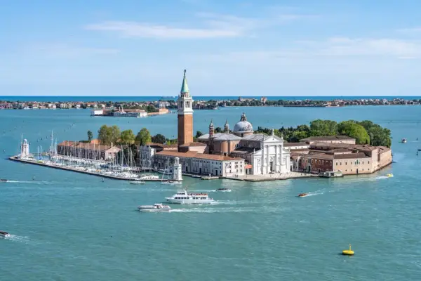 Widok na wyspę San Giorgio Maggiore w Wenecji, z kościołem i dzwonnicą. Wokół pływają łodzie, w tym prom.