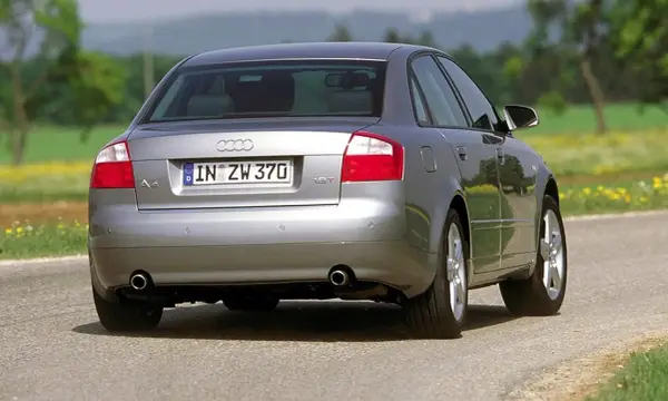 Srebrne Audi B6 sedan z tablicą rejestracyjną IN ZW 370 jedzie po asfaltowej drodze wśród zieleni i żółtych kwiatów.