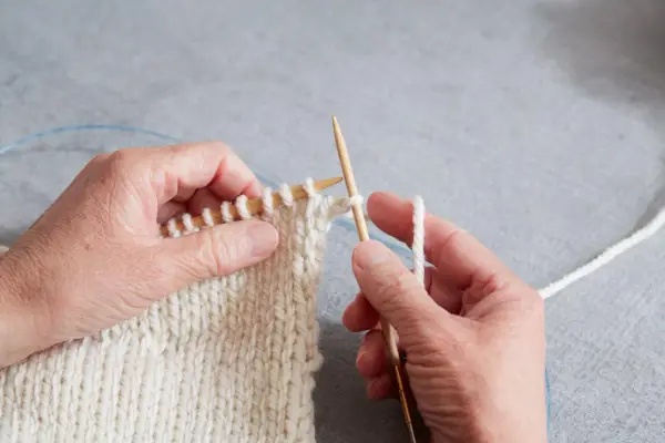 Händer stickar en vit resår med trästickor. En blå kabel syns i bakgrunden.