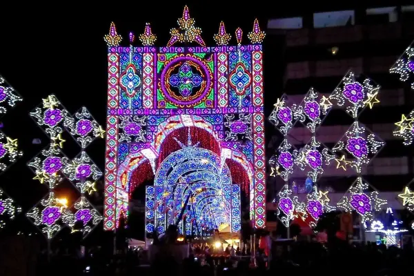 Un tunnel di luci colorate e decorazioni luminose, tipiche delle feste patronali, crea un'atmosfera magica e festosa.
